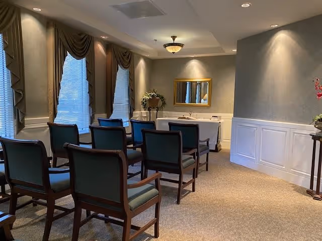 A small meeting or chapel room with two rows of wooden chairs with green cushions facing a white table against the wall. The room has three tall windows with draped curtains, beige walls with white wainscoting, a ceiling light fixture, a gold-framed mirror above the table, and floral arrangements on the table and a side stand.