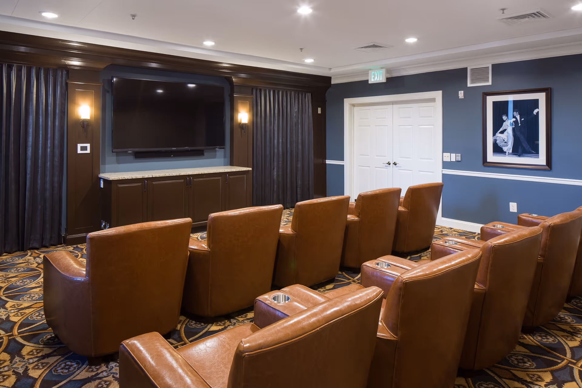 A cozy media room with two rows of brown leather recliner chairs facing a large flat-screen TV mounted on a dark wood cabinet. The room has dark curtains, blue walls with white trim, a patterned carpet, and a framed picture on the wall near a white double door.