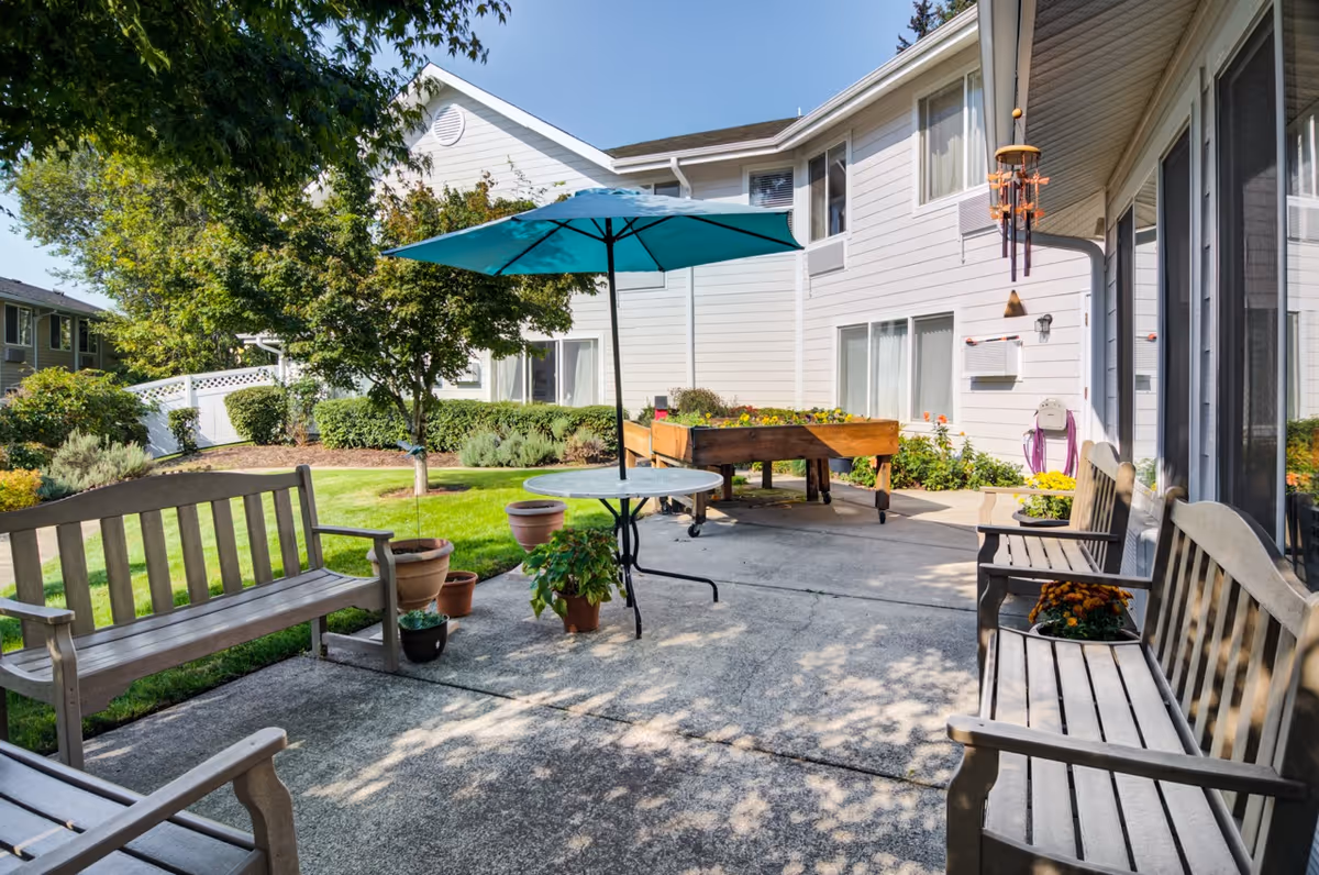 Outdoor patio area at Edgewood Point Senior Living with wooden benches, a round table with a blue umbrella, potted plants, and a raised garden bed. The patio is adjacent to a white building with windows and surrounded by green grass and trees under a clear blue sky.