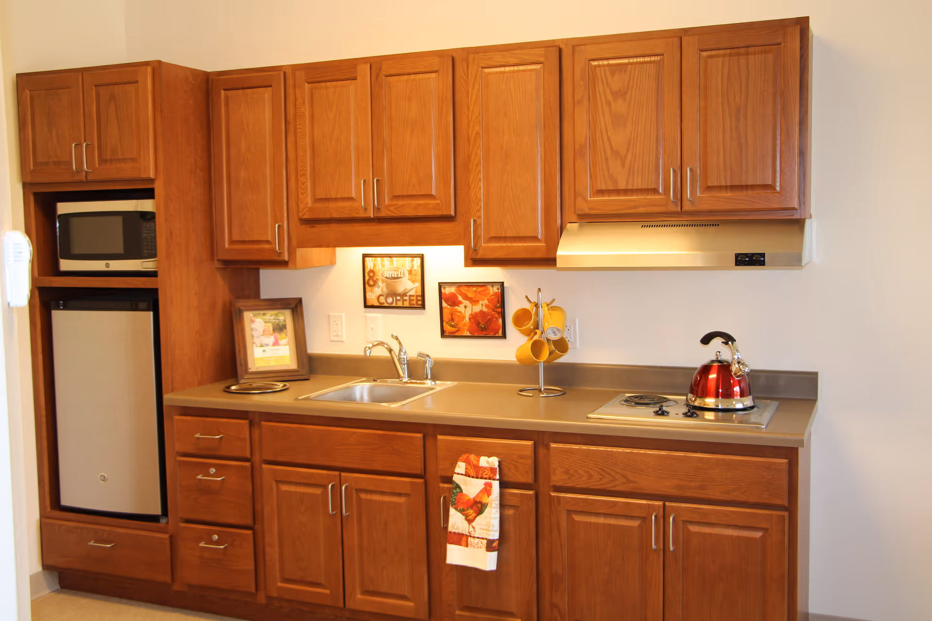 A compact kitchen area with wooden cabinets, a small refrigerator, a microwave, a sink, a stovetop with a red kettle, and a few decorative items including a framed picture, a sign that says 'Wake up & smell the coffee', and a rack holding yellow mugs.