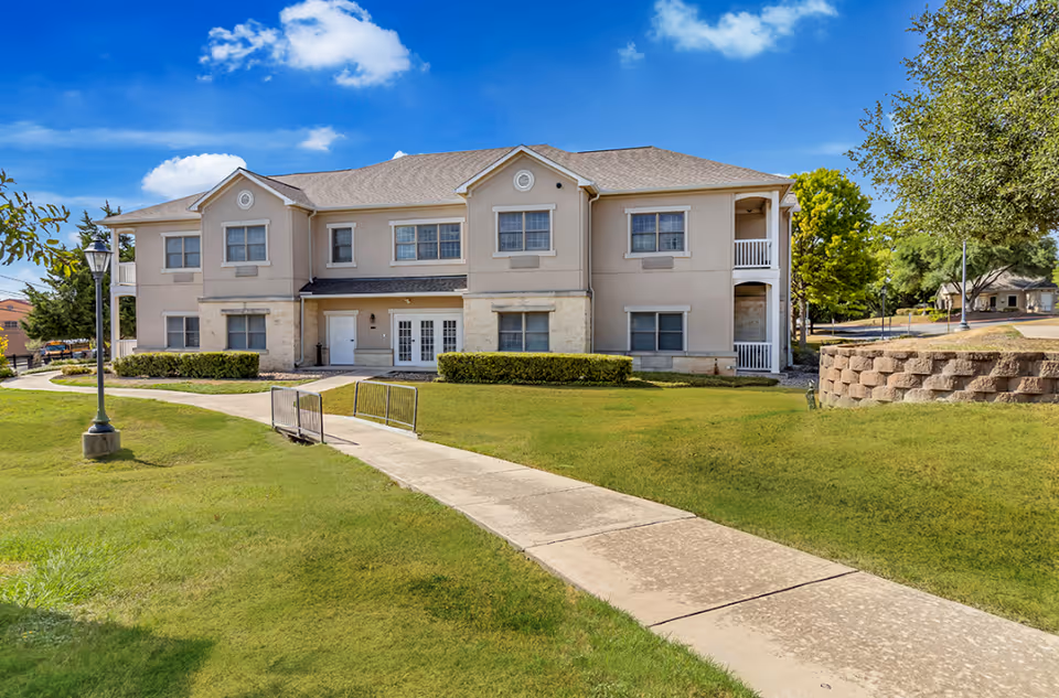 Exterior view of a two-story senior living facility building with beige walls and multiple windows, surrounded by green grass, a paved walkway, and trees under a blue sky with some clouds.