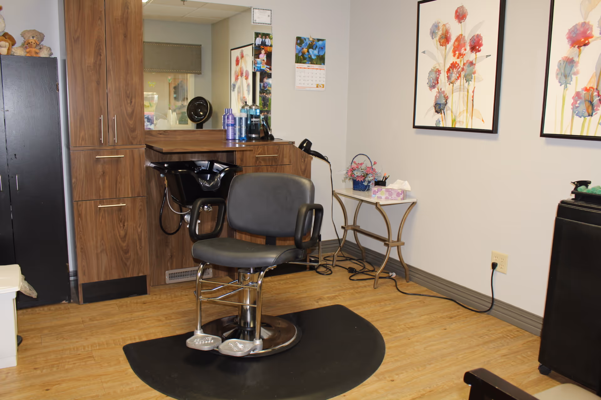 Interior of a hair salon area within a senior living facility featuring a black salon chair on a black mat, a wooden cabinet with hair care products, a black hair washing sink, a small table with a tissue box and a flower arrangement, and two framed floral paintings on the wall.