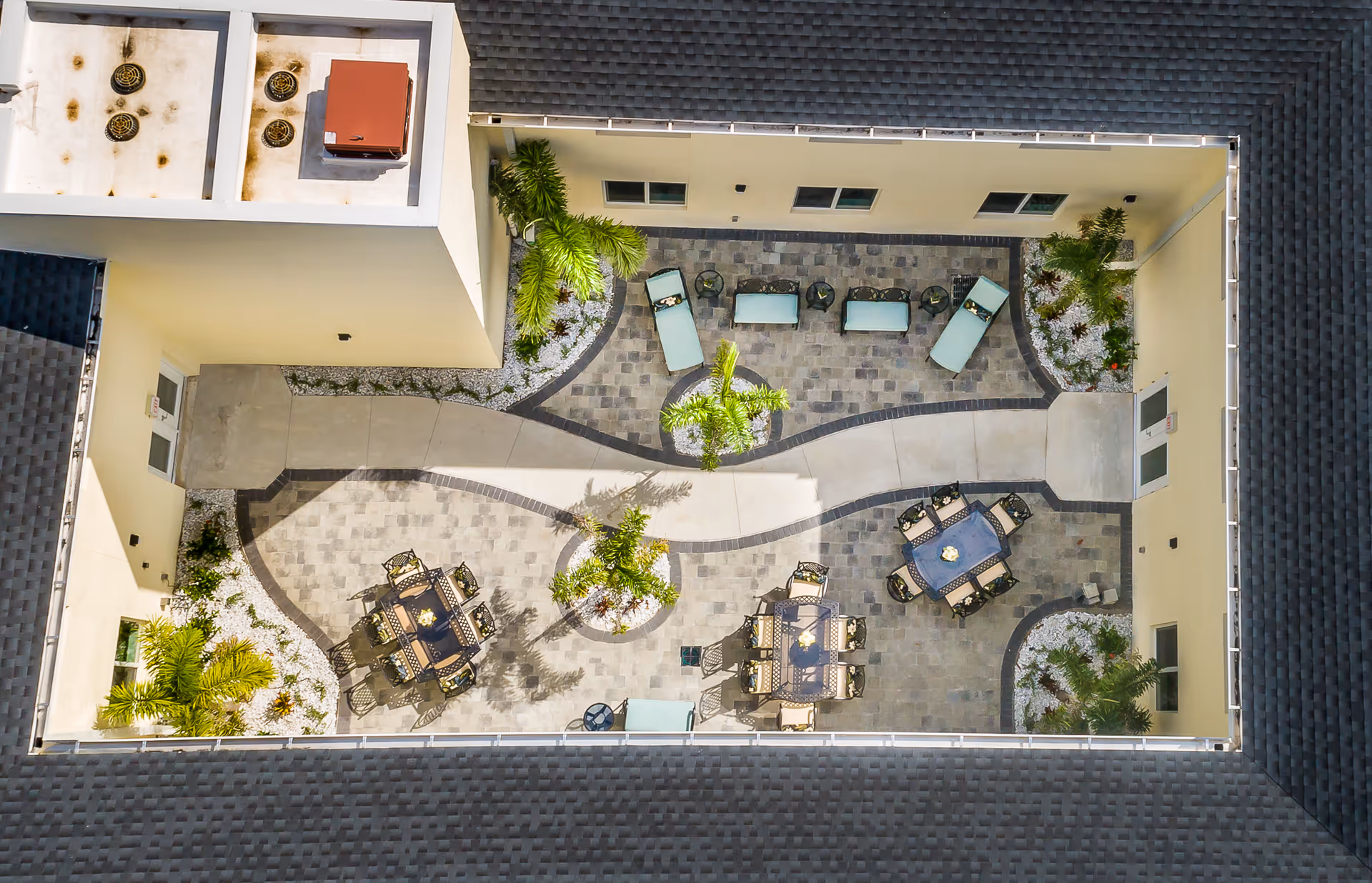 Aerial view of an enclosed rectangular courtyard with paved walkways, tables and chairs, lounge chairs, and planted palm trees.