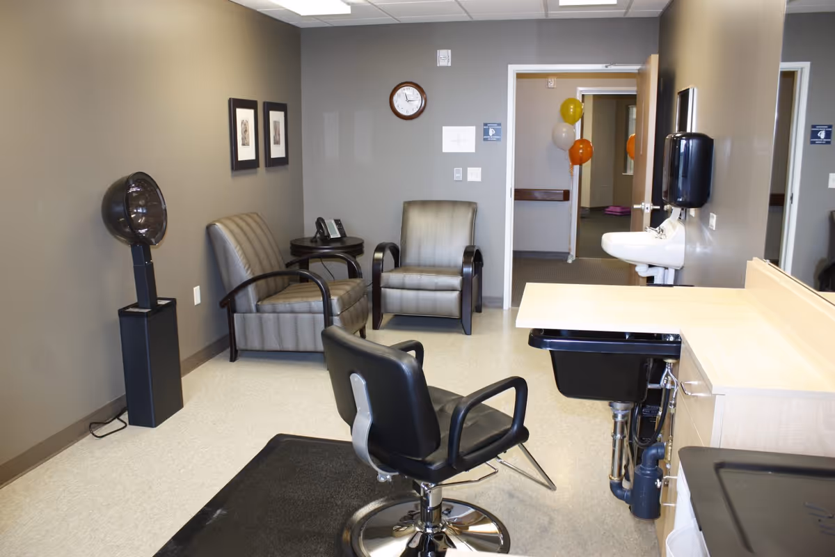 Interior view of a salon area in a senior living facility with a black salon chair on a black mat, a hair dryer on a stand, two armchairs with a small round table and a phone between them, a wall clock, a sink with a soap dispenser, and balloons visible through an open door in the background.