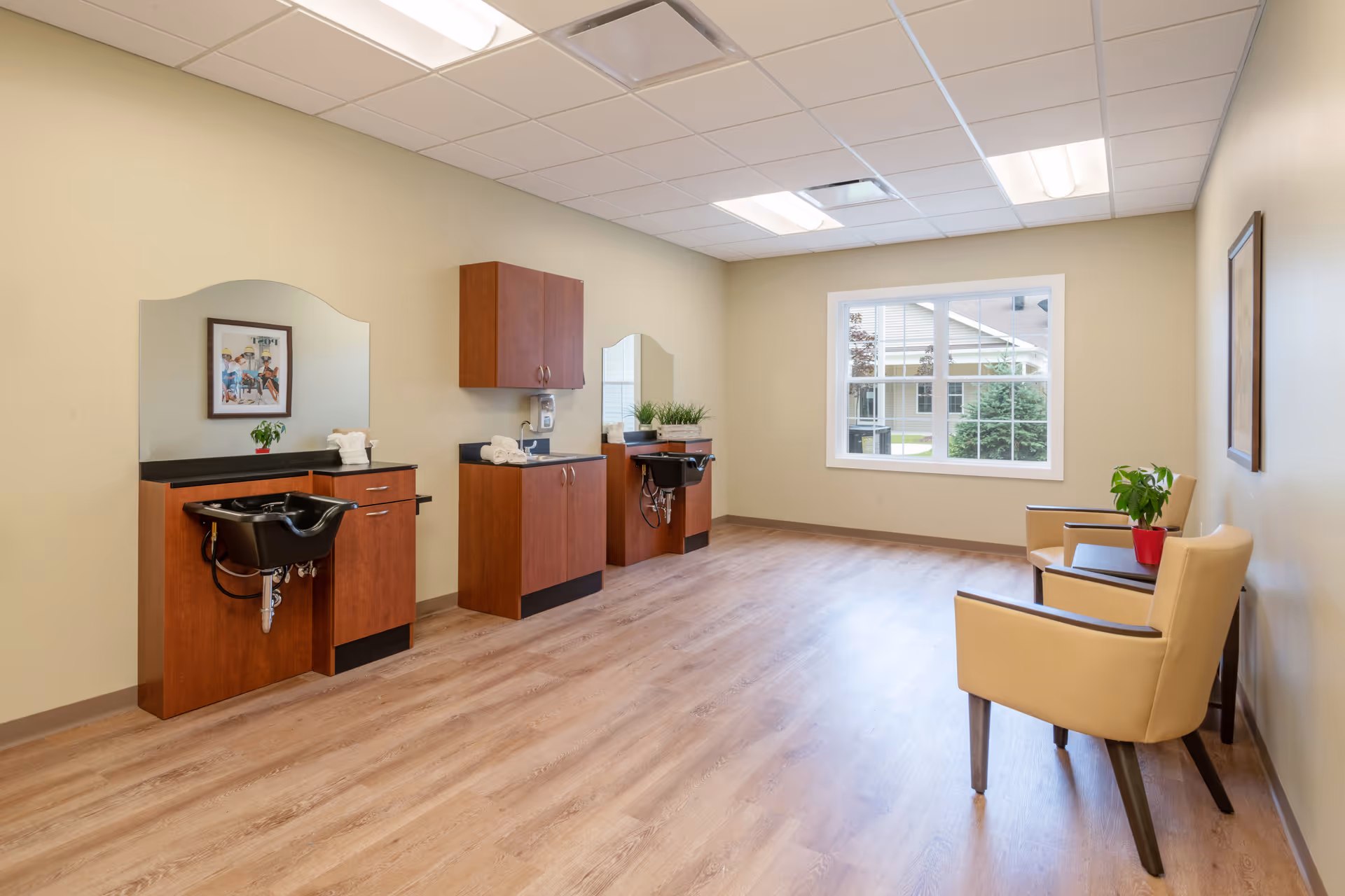 A clean and well-lit hair salon area in a senior living facility with two hair washing stations featuring black sinks and wooden cabinetry. There are two beige chairs with a small table and a potted plant near a large window letting in natural light. The walls are light yellow, and the floor is wood laminate.
