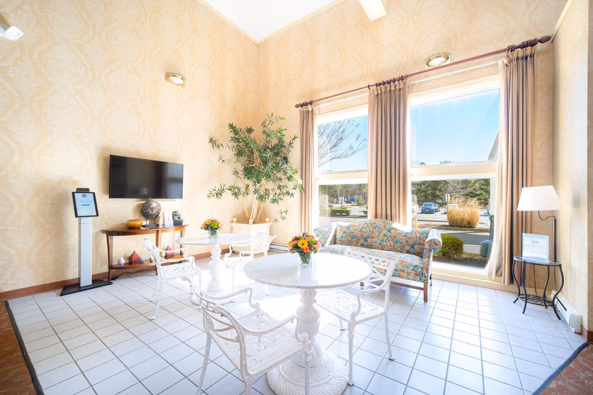 A bright and airy living room area with large windows letting in natural light. The room features a floral-patterned sofa, white metal chairs around a round white table with a small flower arrangement, a flat-screen TV mounted on the wall, a wooden console table with decorative items, a tall green plant, and beige patterned wallpaper.