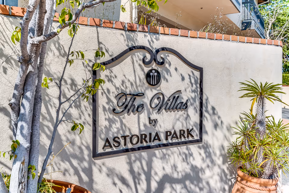 A decorative sign mounted on a beige stucco wall with a brick trim on top, displaying the text 'The Villas by Astoria Park'. There are shadows of tree branches on the wall, a tree trunk on the left, and a potted plant with spiky leaves on the right side of the image.