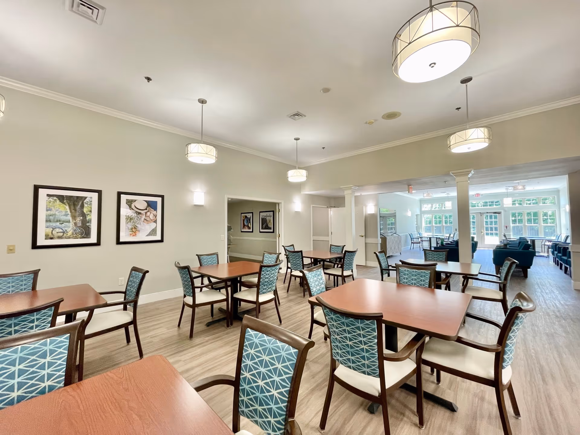 A bright and spacious dining room in a senior living facility with multiple wooden tables and chairs featuring blue patterned upholstery. The room has light-colored walls adorned with framed artwork, modern ceiling lights, and large windows allowing natural light to fill the space. In the background, there is a lounge area with additional seating and large windows.