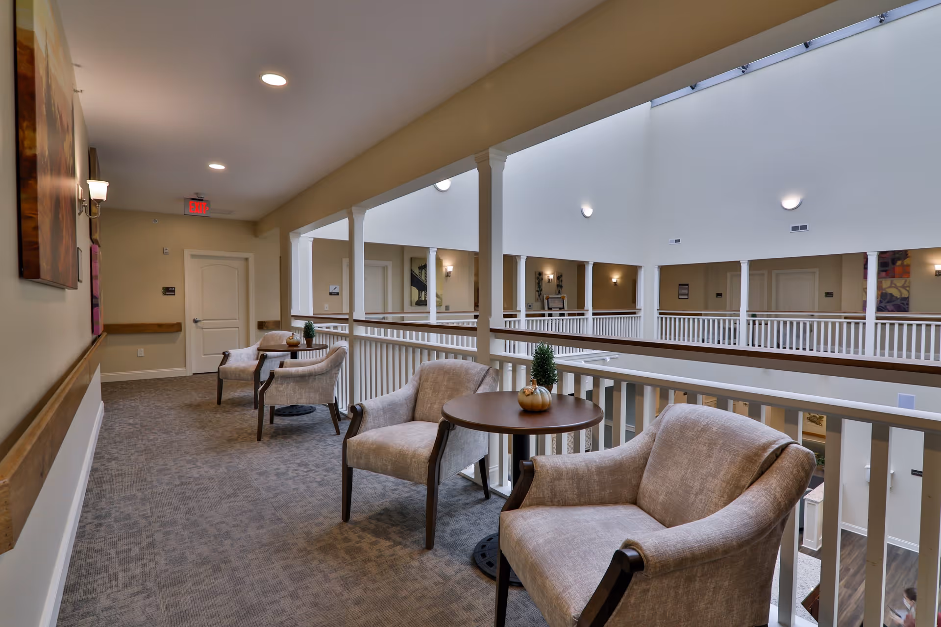 A bright and spacious indoor balcony area in a senior living facility with beige walls and carpeted floor. The area features comfortable beige armchairs paired with small round tables decorated with small plants and a pumpkin. White railings surround the balcony overlooking the lower floor, and multiple doors and wall sconces are visible along the corridor.