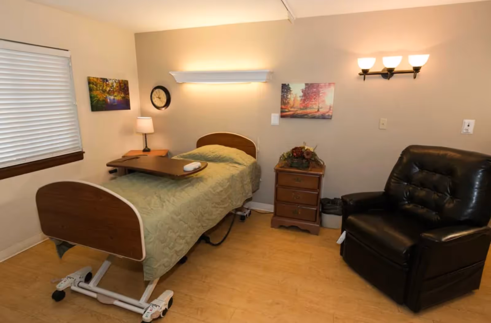 A nursing facility bedroom with a single hospital-style bed covered with a green quilt, a wooden bedside table with a lamp, a wall clock, two framed pictures on the walls, a black recliner chair, and a small wooden dresser with a floral arrangement. The room has light-colored walls and a window with closed blinds.