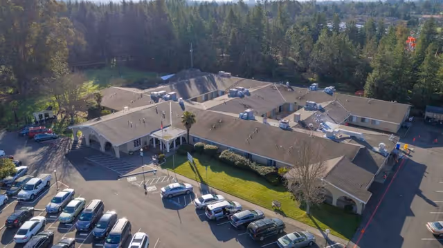 Aerial view of Broadway Villa Post Acute facility showing a single-story building with multiple roof sections surrounded by trees and a parking lot filled with cars.