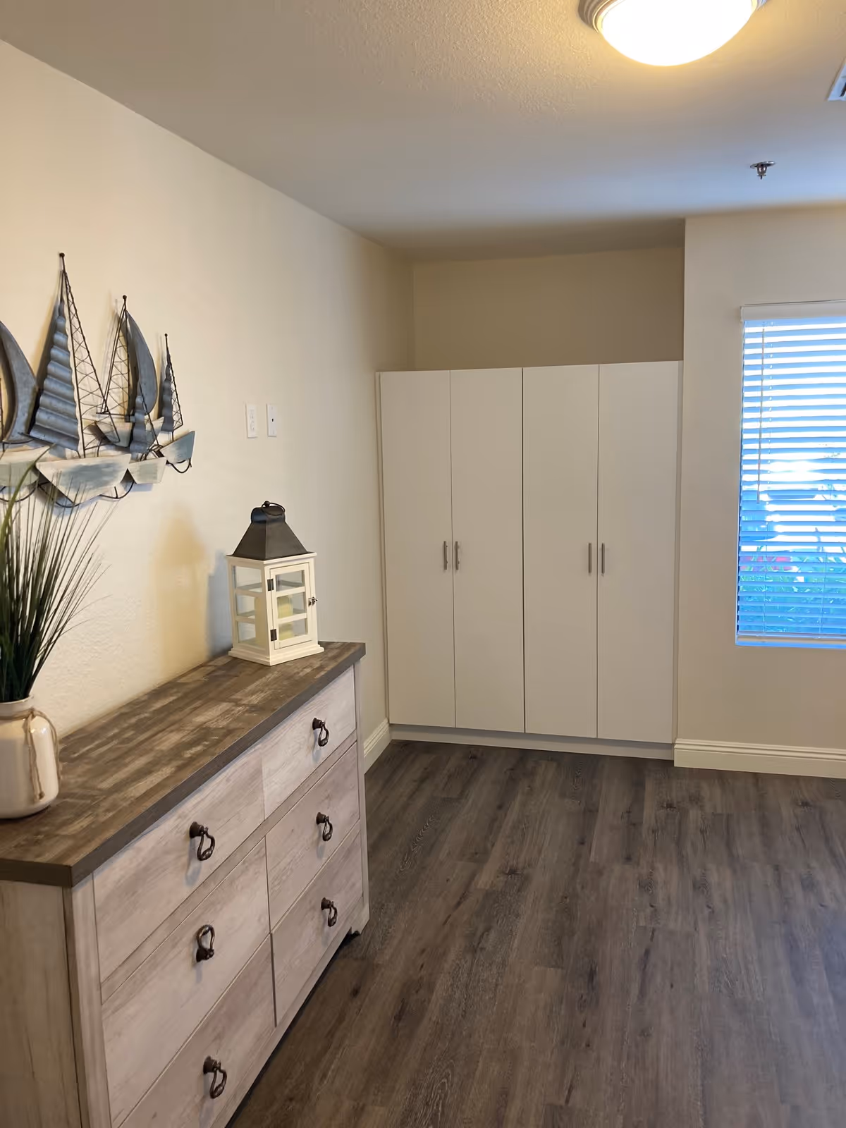 A corner of a room with a wooden dresser on the left side, decorated with a potted plant and a white lantern. Above the dresser is a metal wall art piece depicting three sailboats. On the right side, there is a white cabinet with four doors and a window with white blinds letting in natural light. The floor is covered with dark wood laminate.
