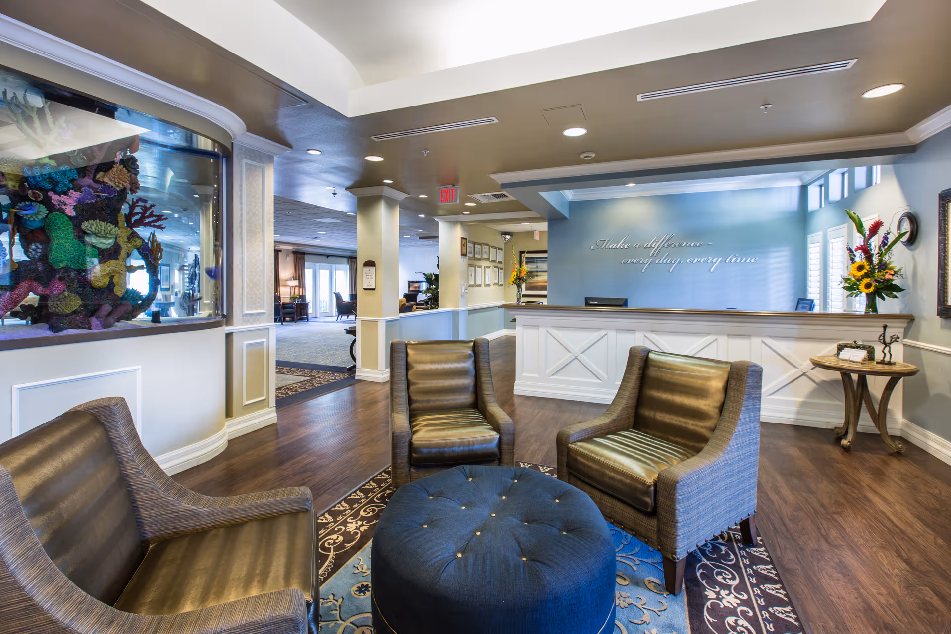 A bright and welcoming senior living facility lobby with a reception desk, three comfortable armchairs arranged around a round ottoman on a patterned rug, a large aquarium built into the wall, and a side table with a flower arrangement. The wall behind the reception desk has the phrase 'Make a difference every day, every time' written on it.
