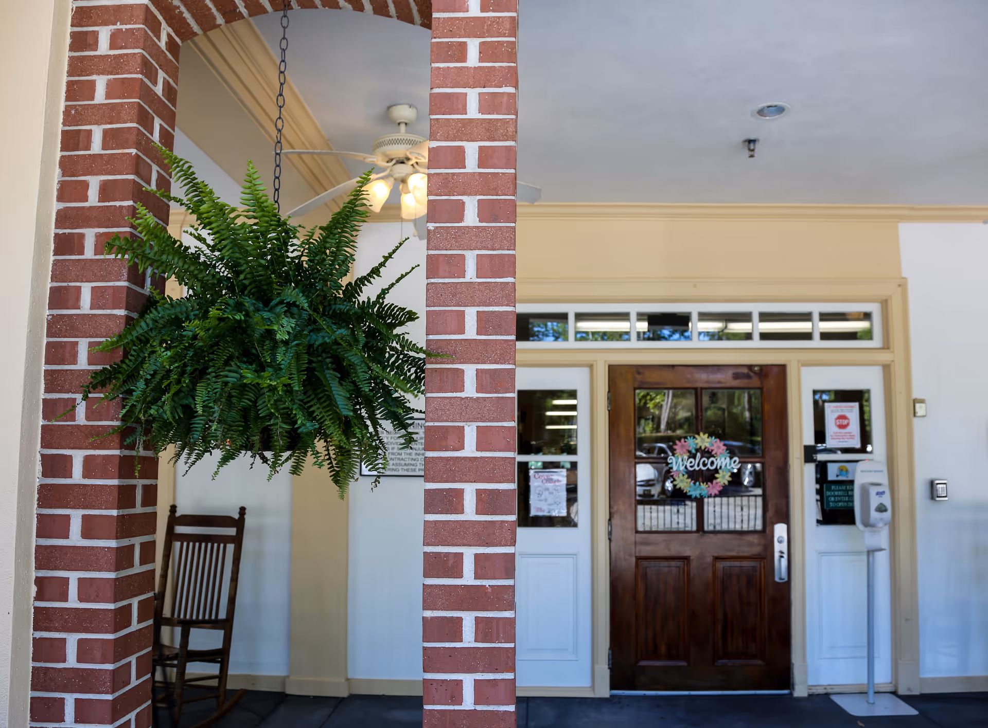 Entrance area of a facility with a wooden door featuring a colorful 'Welcome' sign. The door is flanked by white panels with windows above. A hanging green fern plant is suspended from a brick archway on the left side, and a wooden rocking chair is partially visible behind the arch. A hand sanitizer dispenser is mounted on the right side near the door.