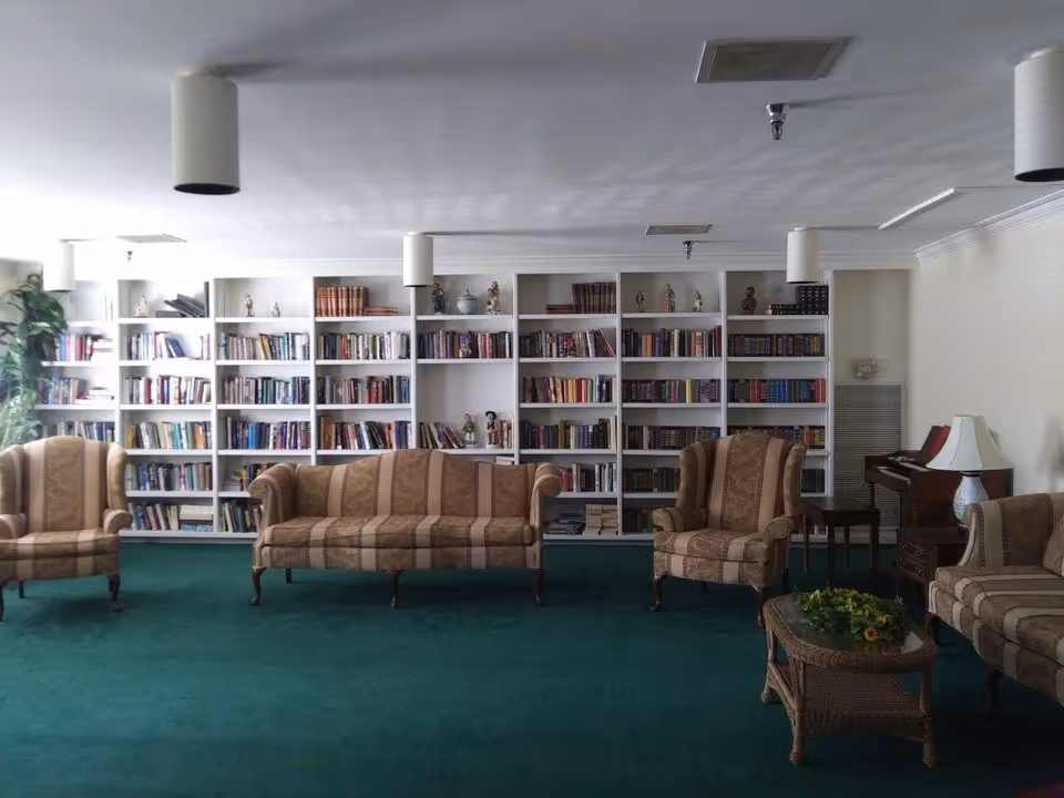 A cozy lounge with bookshelves along the back wall and striped sofas and armchairs arranged on a green carpet.