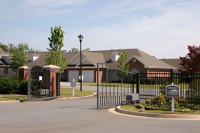 Gated driveway entrance leading to single-story senior living buildings with lawns and trees.