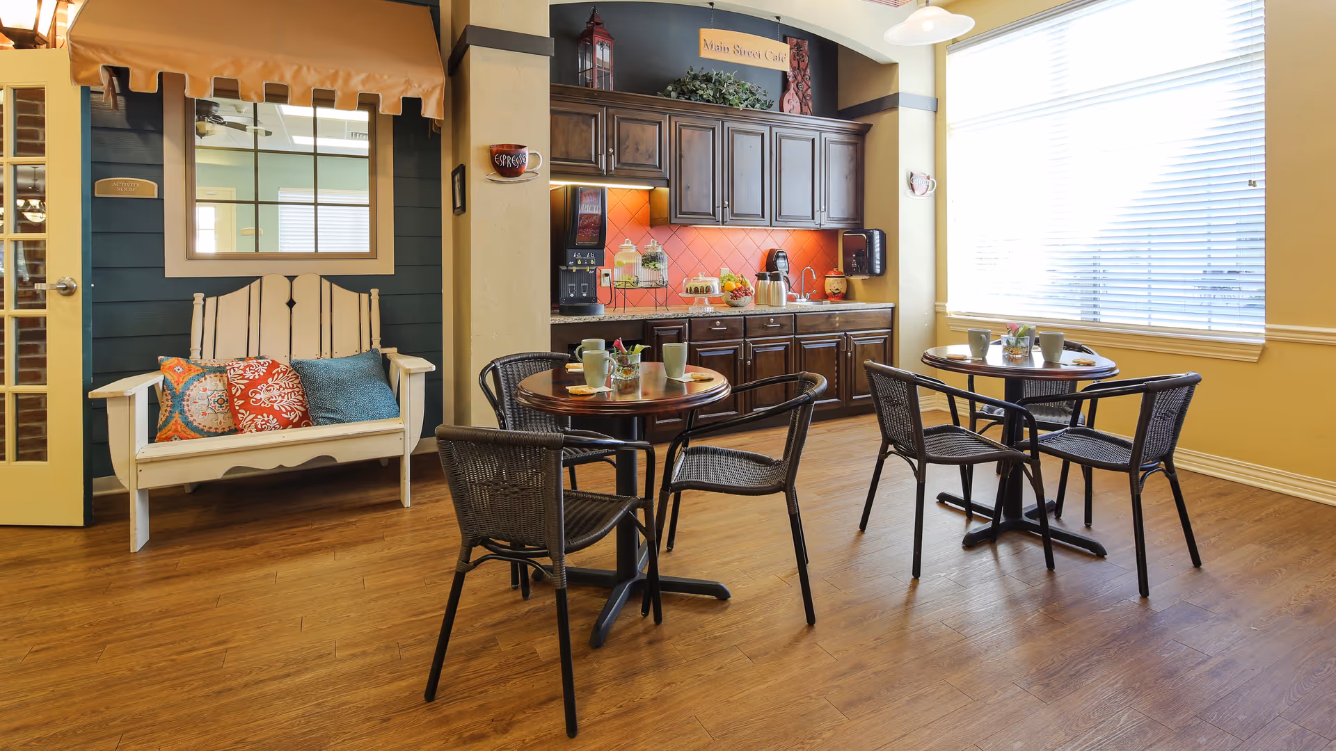 A cozy cafe-style seating area with two round tables, each surrounded by four black wicker chairs. The tables have cups and small flower arrangements. Behind the tables is a wooden cabinet with a coffee machine, snacks, and a sign that reads 'Main Street Cafe'. To the left, there is a white wooden bench with colorful cushions against a dark green wall with a window and a yellow door. The room has wooden flooring and large windows with blinds letting in natural light.