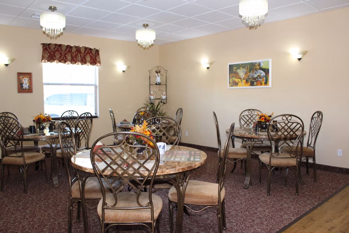 Dining room with several round marble-top tables and metal chairs, floral centerpieces, wall art, and chandelier-style ceiling lights.