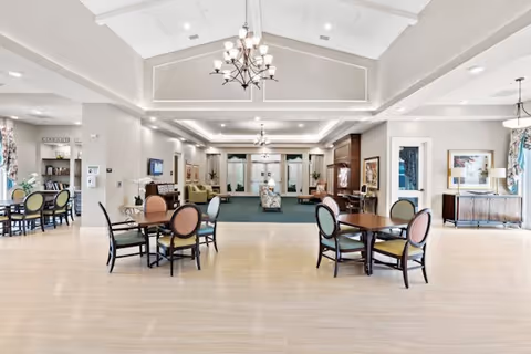 Spacious and well-lit common area in a senior living facility featuring multiple round tables with chairs, a seating area with sofas and armchairs, chandeliers hanging from a high ceiling, and large windows with curtains allowing natural light to fill the room.