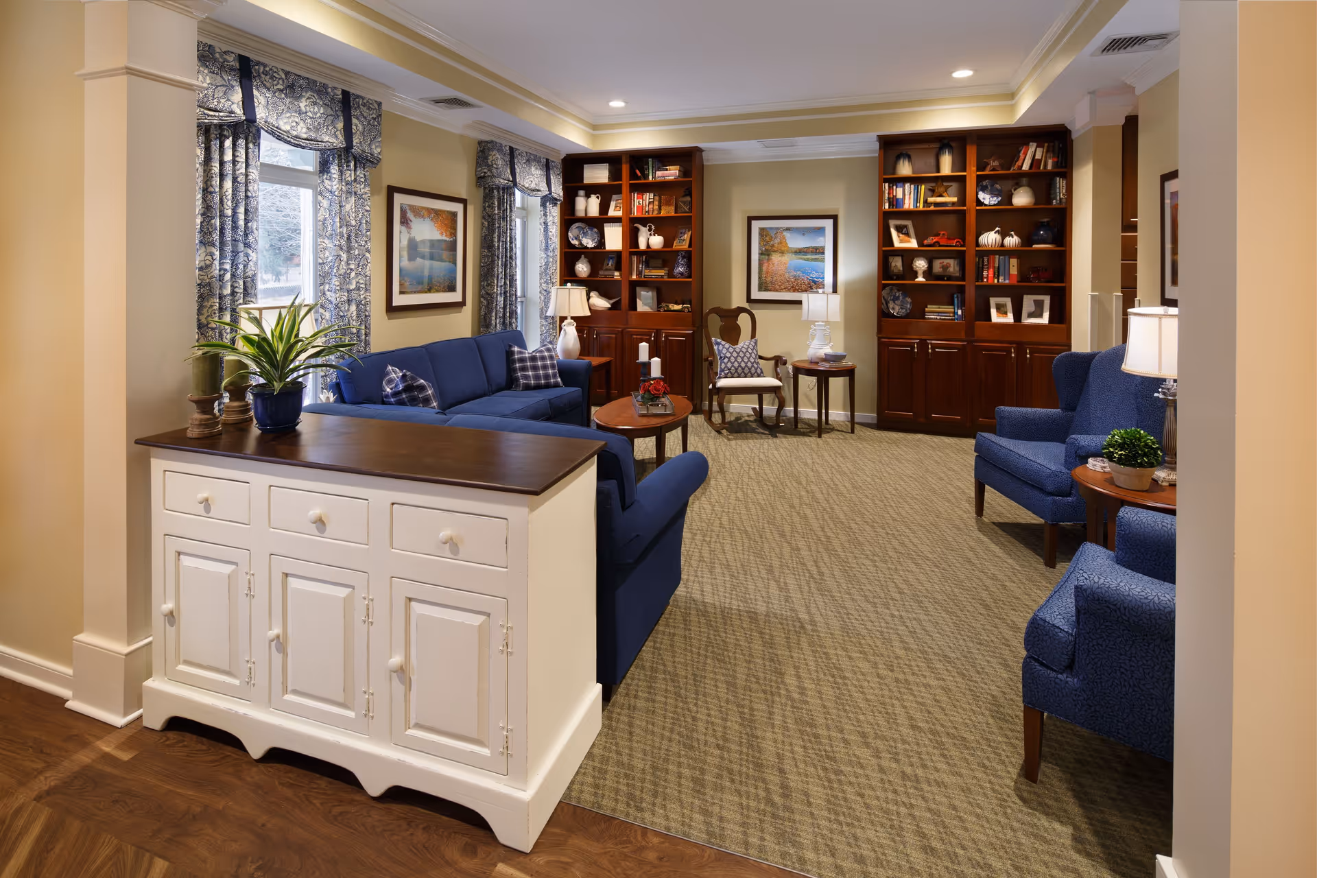 A cozy living room with blue upholstered sofas and armchairs arranged around a wooden coffee table. The room features two large wooden bookshelves filled with books and decorative items, framed artwork on the walls, and windows with patterned blue curtains. A white sideboard with drawers and cabinets is positioned near the entrance, topped with a potted plant and decorative items. The floor is carpeted in the seating area and hardwood near the sideboard, with warm lighting from table lamps.