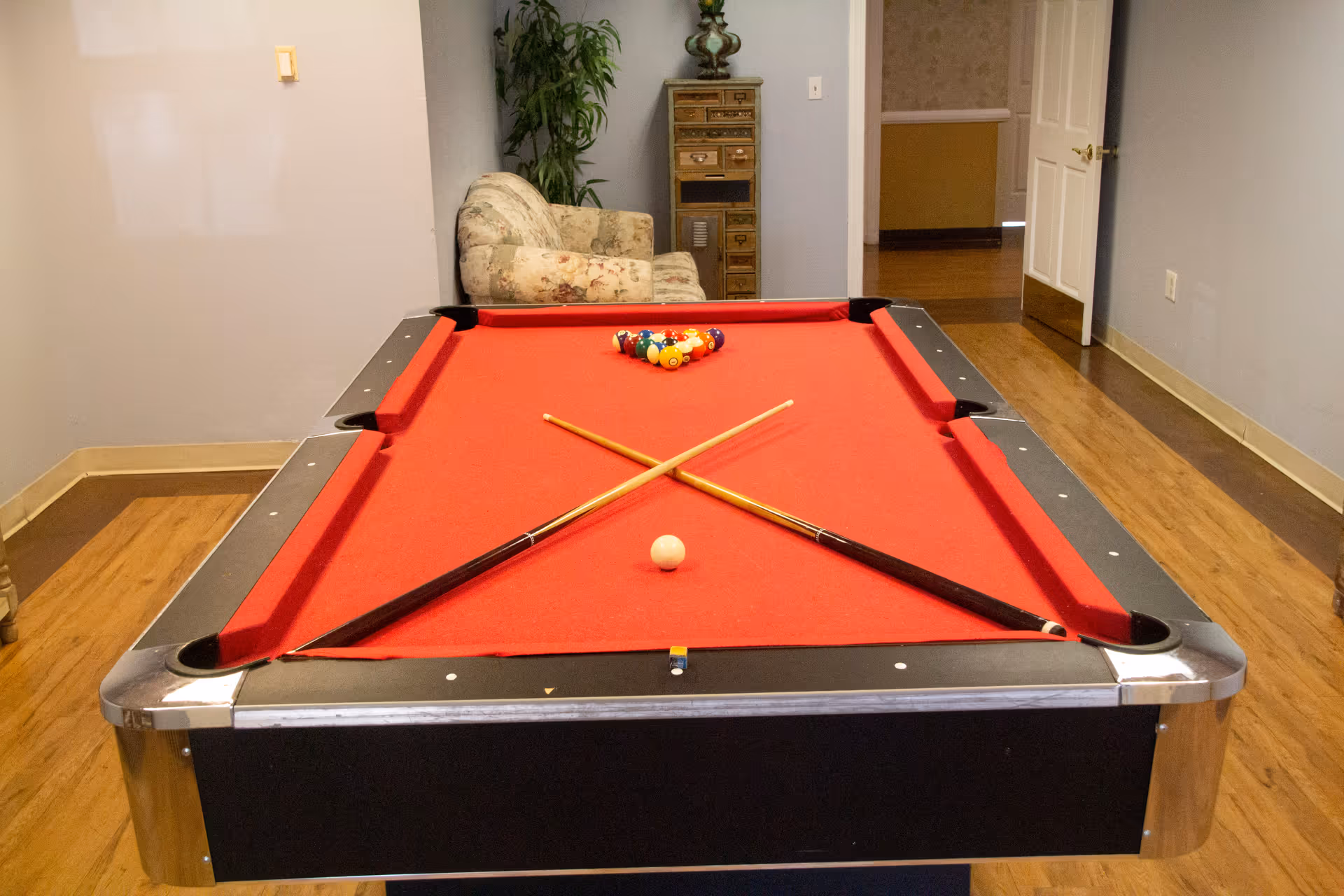A room with a red felt pool table with two crossed pool cues and a white cue ball in the foreground. Behind the pool table is a floral patterned armchair, a tall green plant, and a small wooden cabinet with multiple drawers. The room has light-colored walls and wooden flooring, with an open door leading to another area.