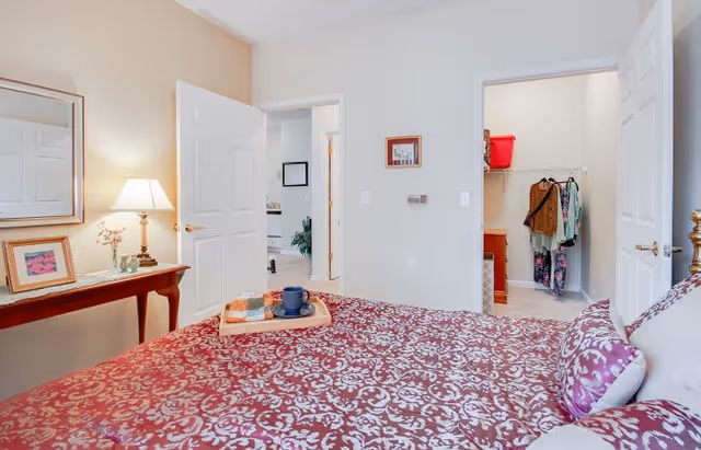 A bedroom with a bed covered in a red and white patterned bedspread and matching pillows. A wooden side table with a lamp, framed pictures, and a small vase with flowers is next to the bed. Two open doors reveal a walk-in closet with clothes hanging and another room with a plant and framed picture on the wall.