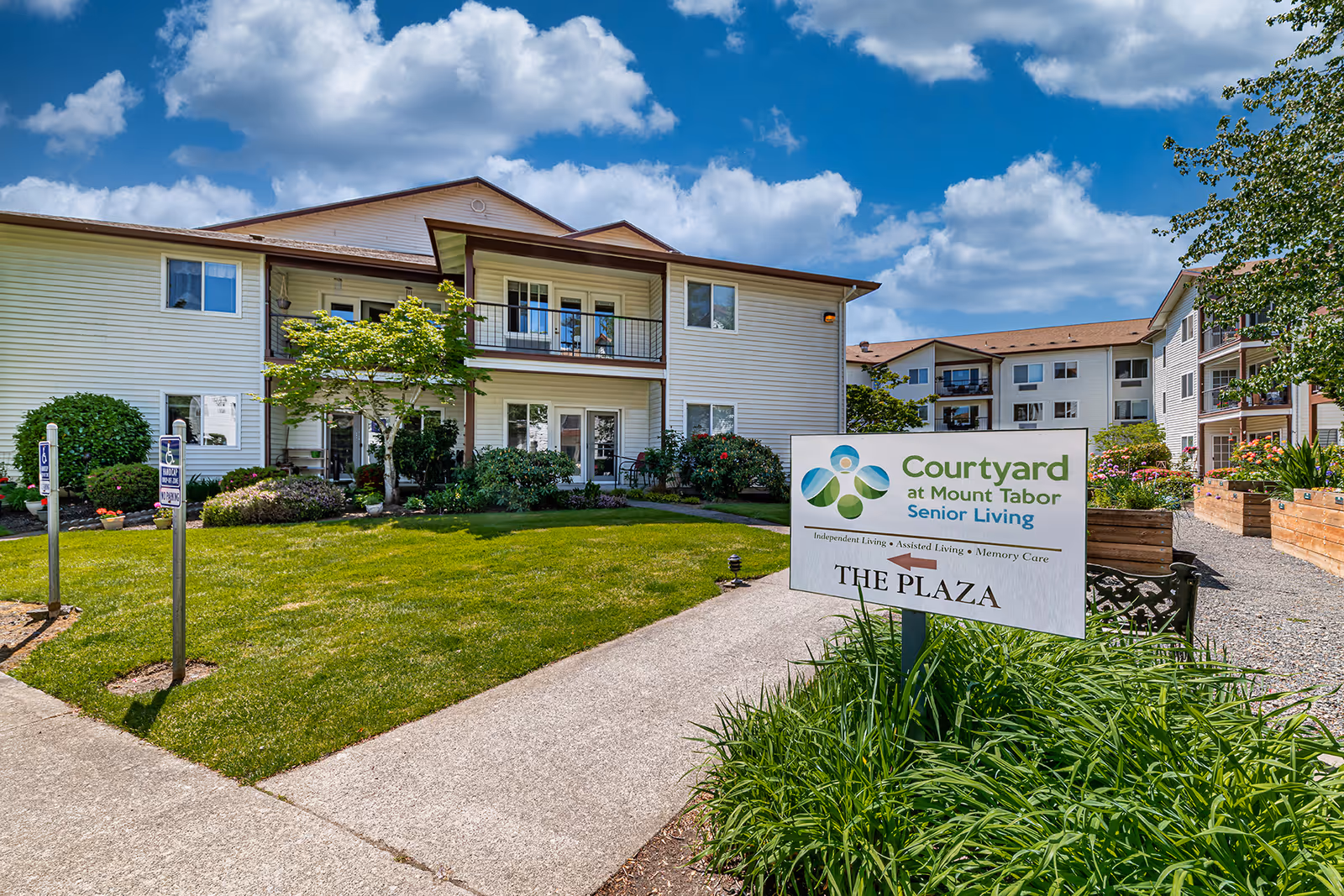 Exterior view of Courtyard at Mount Tabor Senior Living facility showing a two-story building with balconies, well-maintained green lawn, bushes, and a clear blue sky with clouds. A sign in the foreground indicates directions to The Plaza and mentions Independent Living, Assisted Living, and Memory Care.