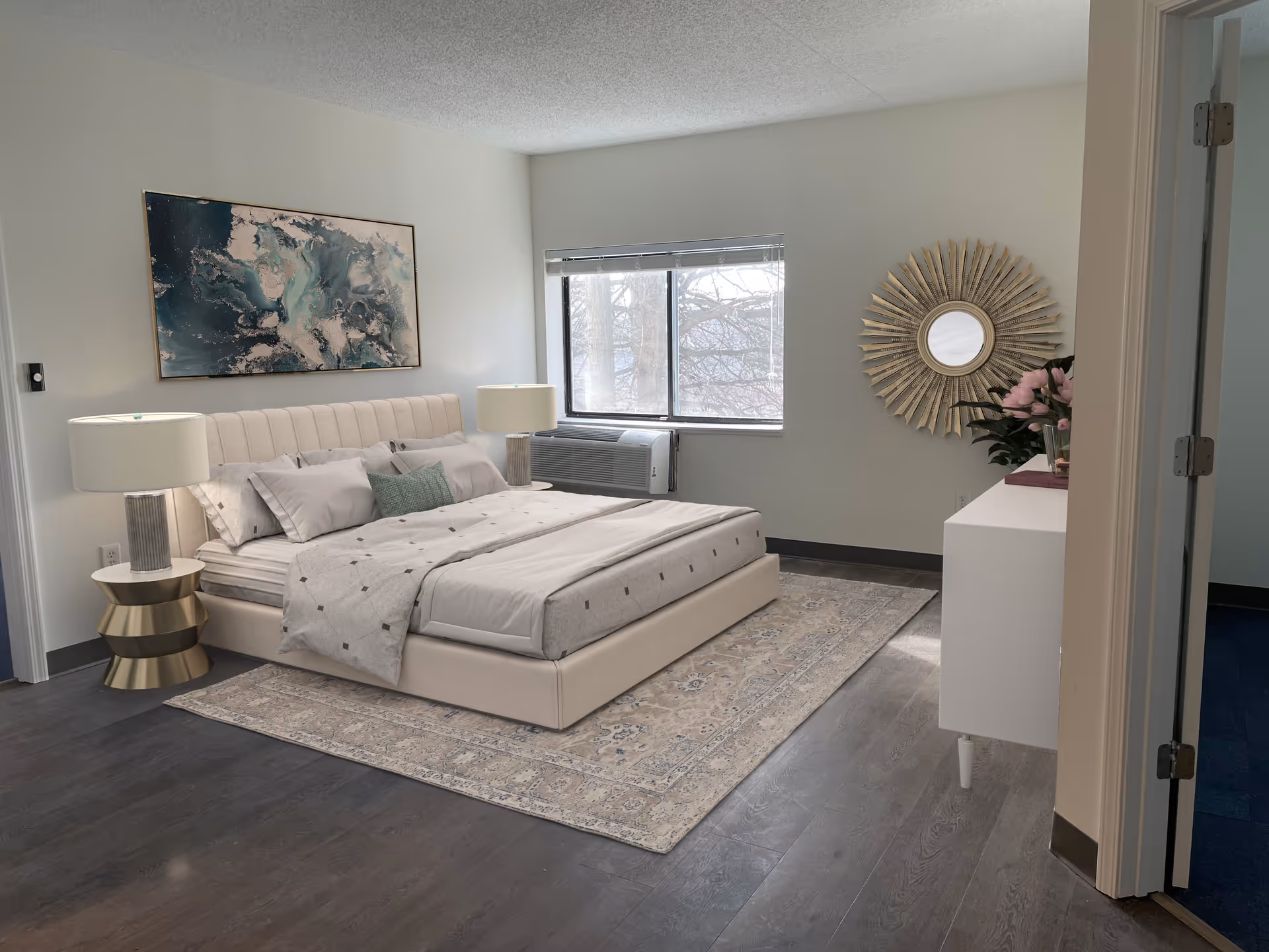 A modern bedroom with a large beige upholstered bed with white and gray bedding, two matching bedside lamps on gold and white nightstands, a large abstract painting above the bed, a window with blinds, a white dresser with a decorative sunburst mirror above it, and a vase with pink flowers. The room has dark wood flooring and light-colored walls.