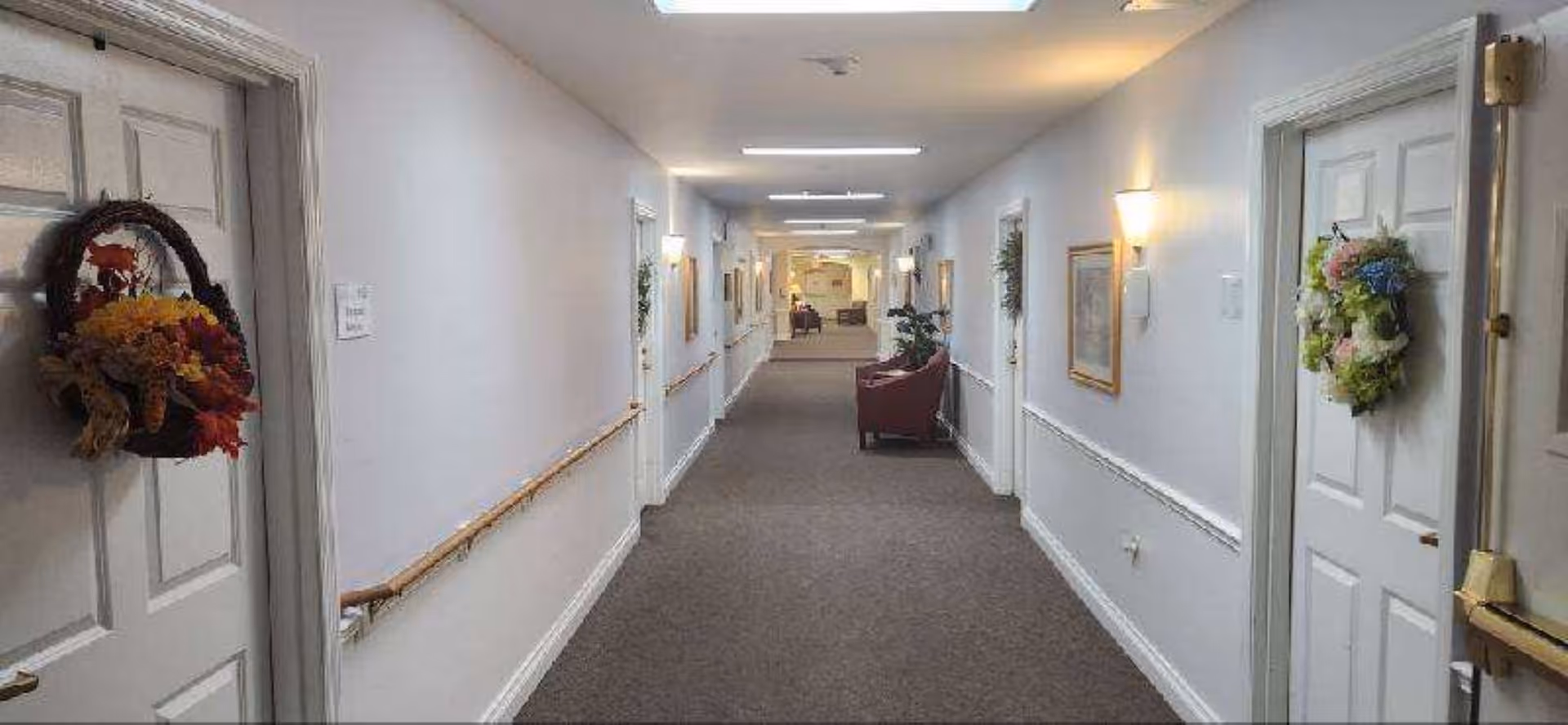 A long, well-lit hallway in a senior living facility with white walls, brown carpet, handrails on both sides, and doors decorated with floral wreaths. There are framed pictures and wall sconces along the walls, and chairs and plants are visible at the far end of the hallway.
