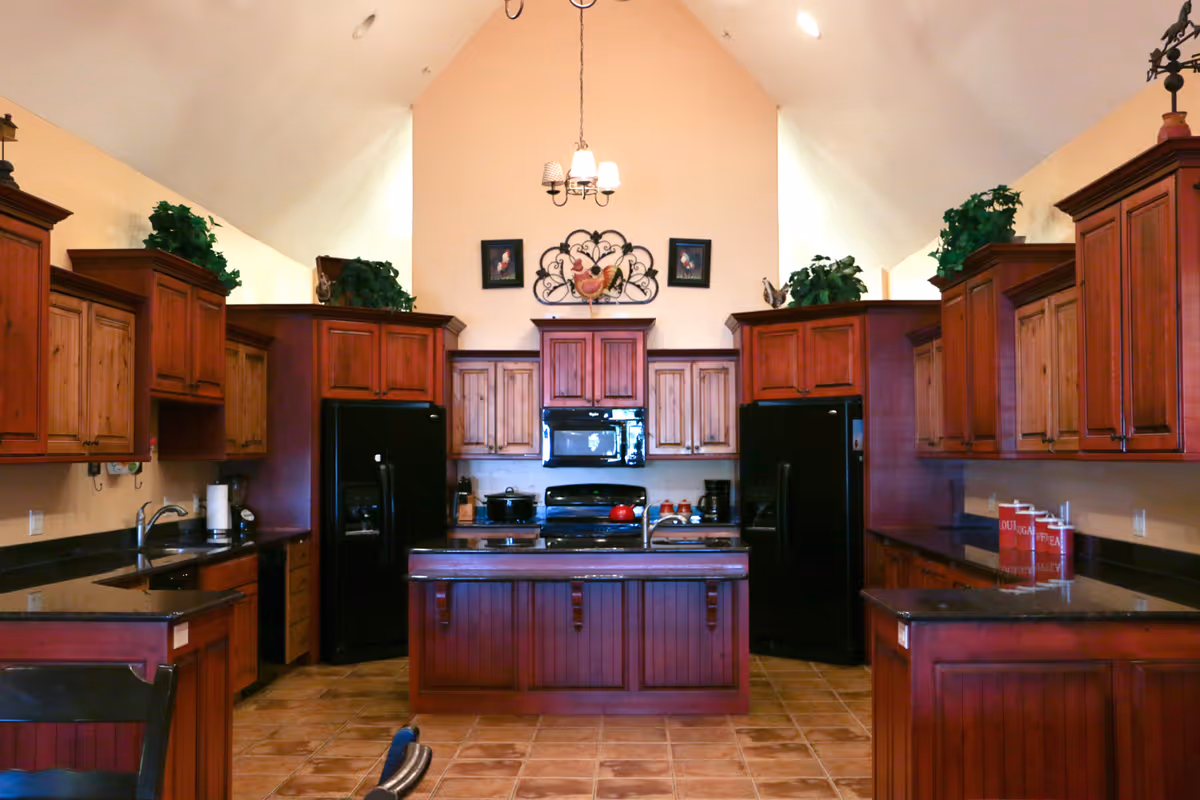A spacious kitchen with high vaulted ceilings, featuring dark wood cabinets and black appliances including two refrigerators, a microwave, and a stove. There is a central island with a sink, granite countertops, and decorative plants on top of the cabinets. The walls are painted a light beige color and there is a chandelier hanging from the ceiling. The floor is tiled with a warm brown pattern.