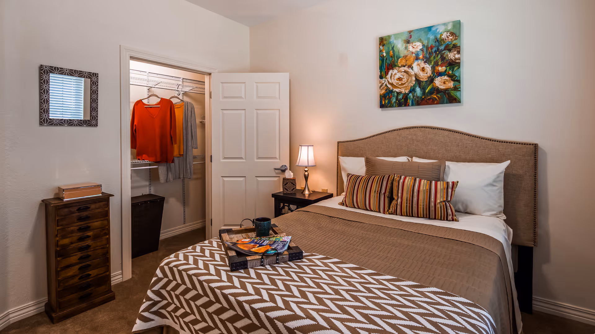 A cozy bedroom with a neatly made bed featuring a beige quilt and striped pillows. A colorful floral painting hangs above the headboard. To the left, there is an open door revealing a walk-in closet with clothes hanging inside. A small wooden dresser with drawers and a decorative mirror are on the left wall. A bedside table with a lamp is next to the bed.