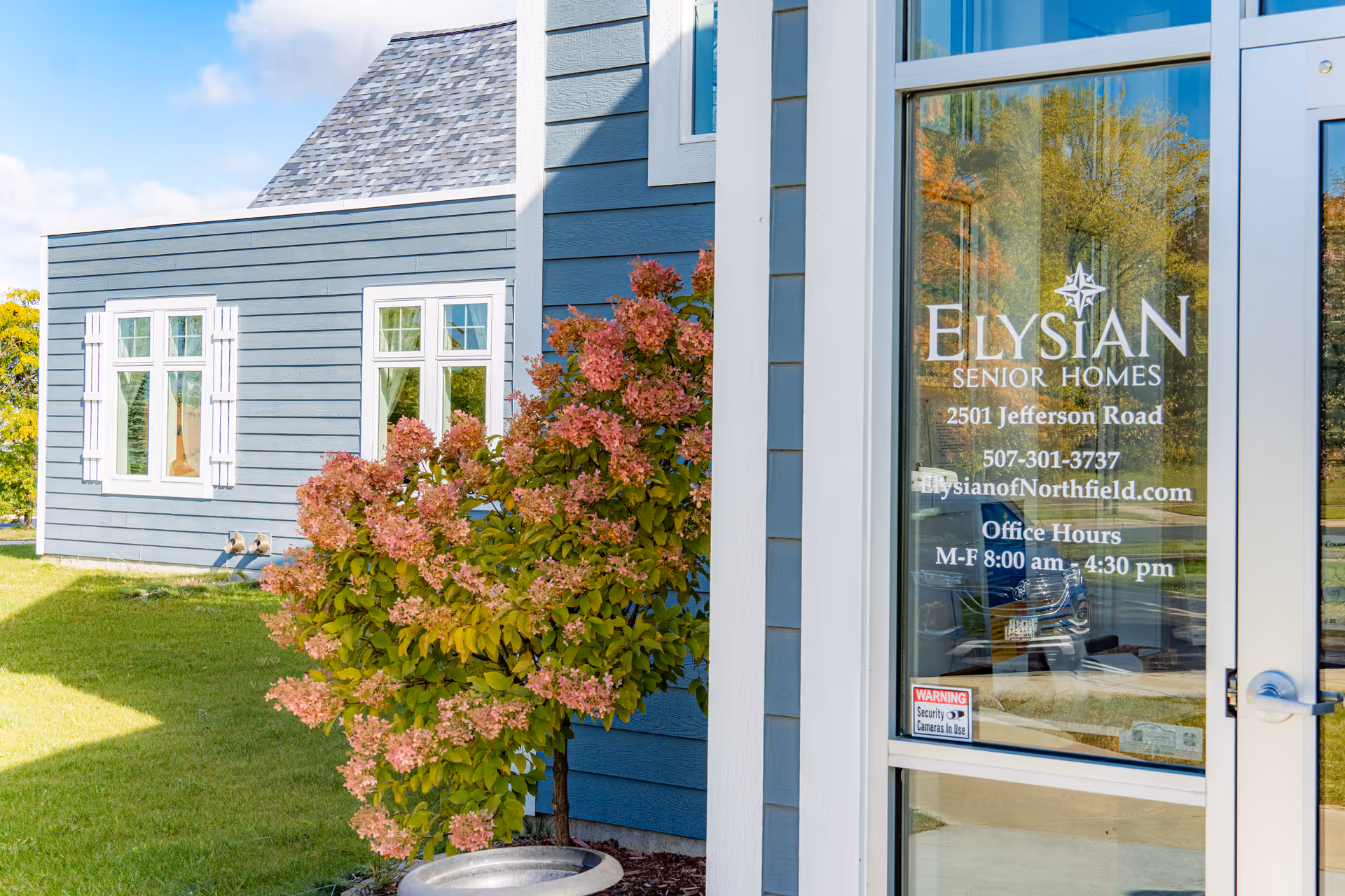 Glass entry door with Elysian Senior Homes signage beside a blue-sided building and a flowering shrub.