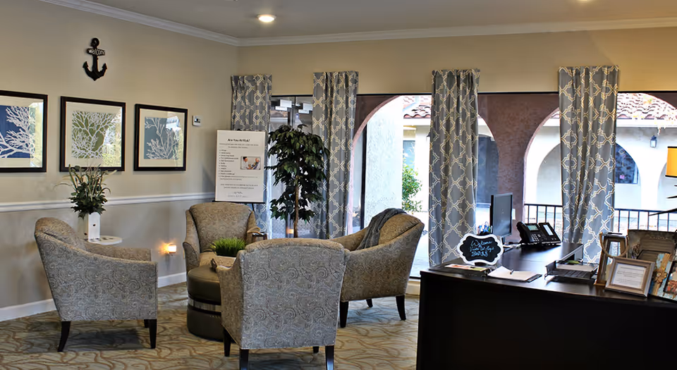 A cozy seating area in a memory care facility with four patterned armchairs arranged around a round ottoman. The room has large windows with patterned curtains, framed artwork on the wall, a potted plant, and a desk with office supplies and a small chalkboard sign.