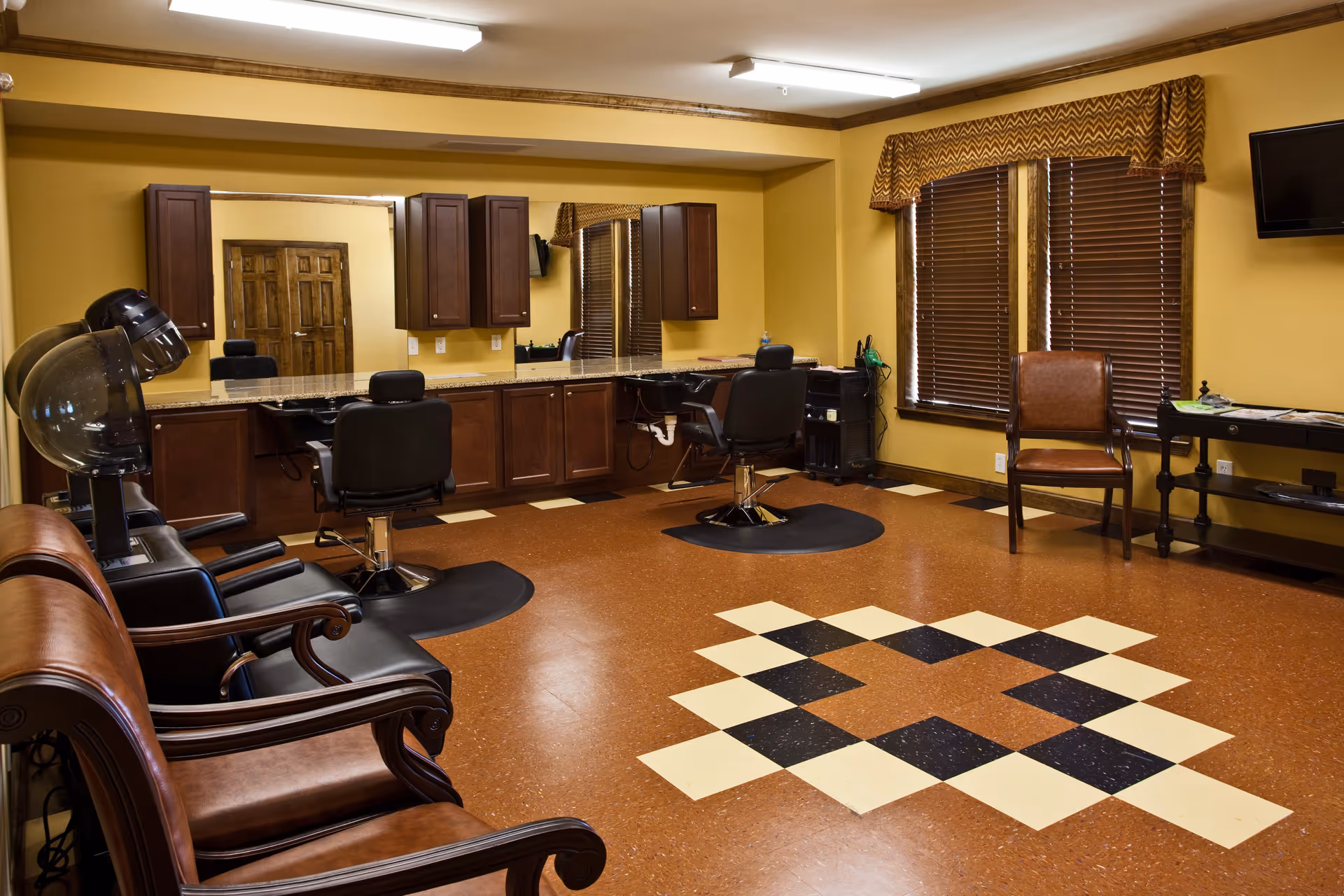 Interior view of a senior living facility's hair salon with brown leather chairs, two black salon chairs in front of a large mirror, hair dryers, wooden cabinets, and a patterned floor design. The room has yellow walls, two windows with brown blinds and valances, and a wall-mounted TV.
