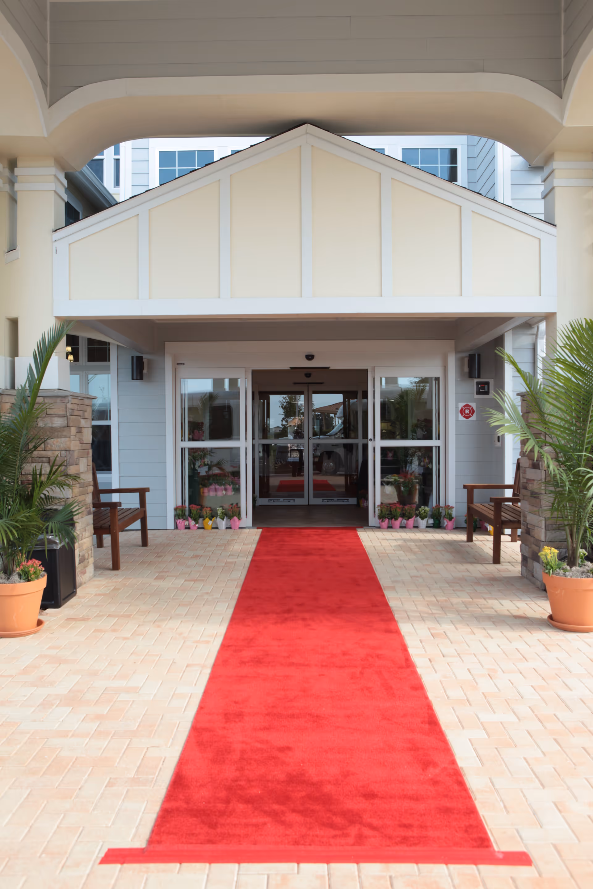 Entrance of The Carriage House Gracious Retirement Living facility with a red carpet leading to automatic glass doors. The entrance is framed by a covered porch with potted plants and wooden benches on either side.