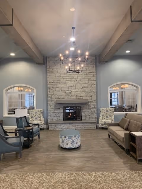 A cozy living room area with a stone fireplace centered on the back wall. Two patterned armchairs are placed on either side of the fireplace, with a round ottoman in front. There are additional seating options including a sofa and chairs arranged around the room. The walls are painted light blue, and two windows with white trim flank the fireplace. A modern chandelier hangs from the ceiling, and wooden beams run across the ceiling.
