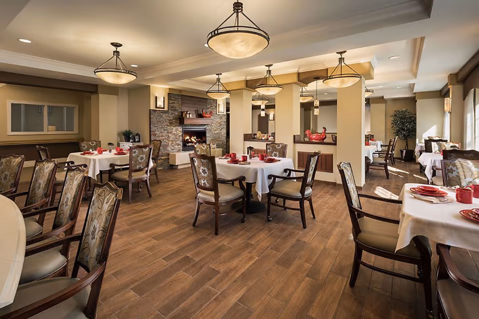 A spacious dining room with multiple round tables covered with white tablecloths, each set with red plates, cups, and utensils. The room features wooden chairs with floral upholstery, a wooden floor, warm lighting from ceiling fixtures, and a stone fireplace in the background. There are decorative elements such as plants and red ceramic roosters on shelves.