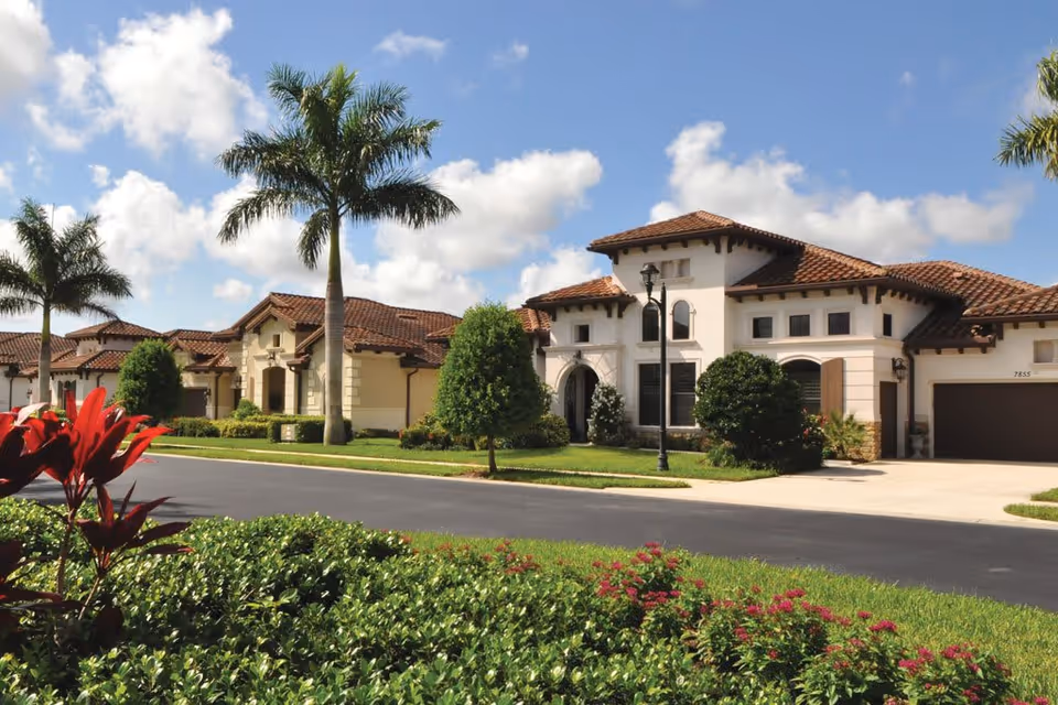 Exterior view of a senior living facility with Mediterranean-style architecture, featuring beige stucco walls, red tile roofs, palm trees, and well-maintained landscaping including green bushes and red flowers under a partly cloudy blue sky.