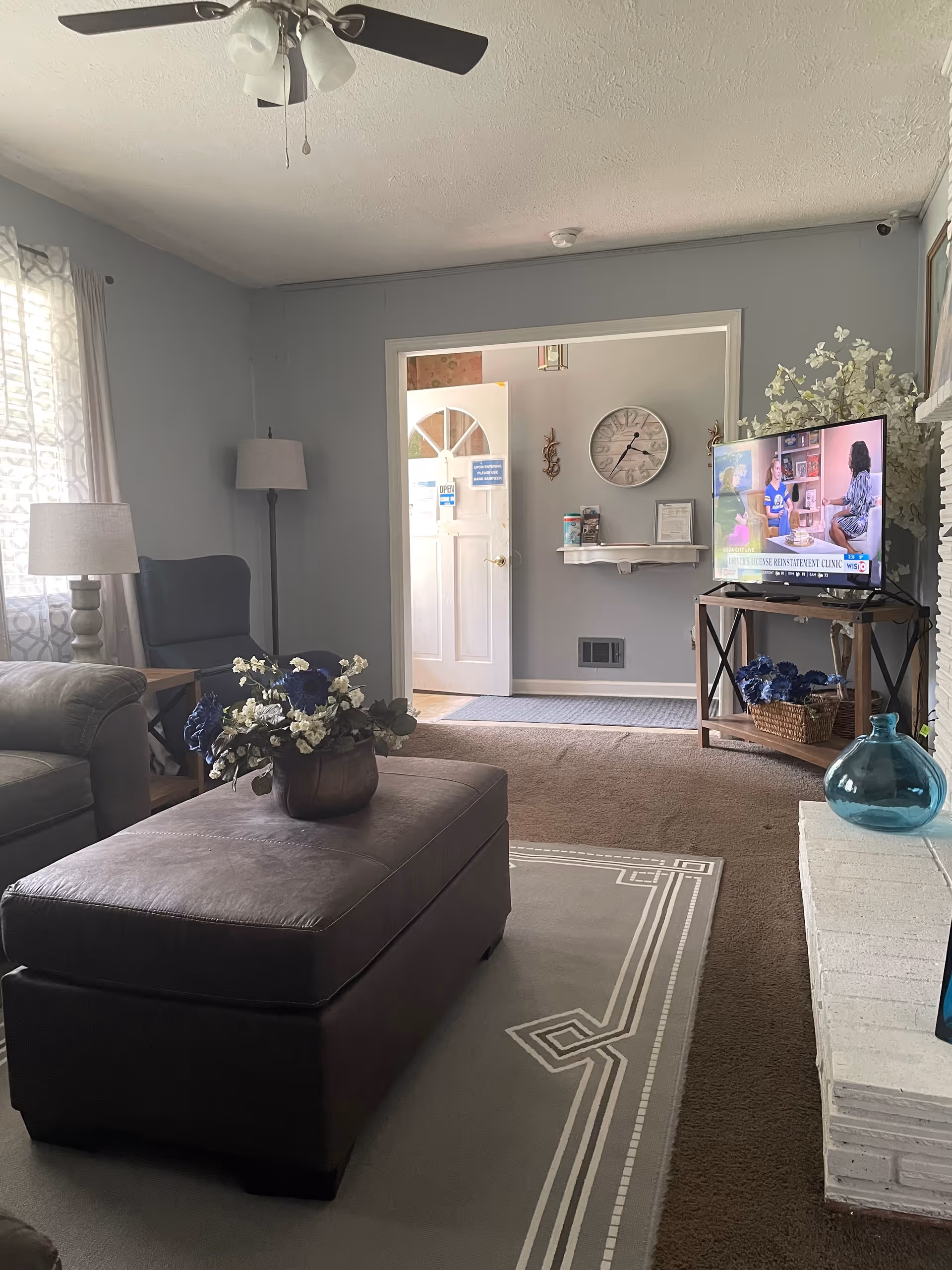 A cozy living room with a ceiling fan and light fixture, a large ottoman with a flower arrangement on top, a gray armchair and floor lamp near a window with patterned curtains. A TV on a stand shows a news program, and a white brick fireplace with a blue glass vase is visible. The room opens into an entryway with a white door, a wall clock, and a small shelf with items on it.