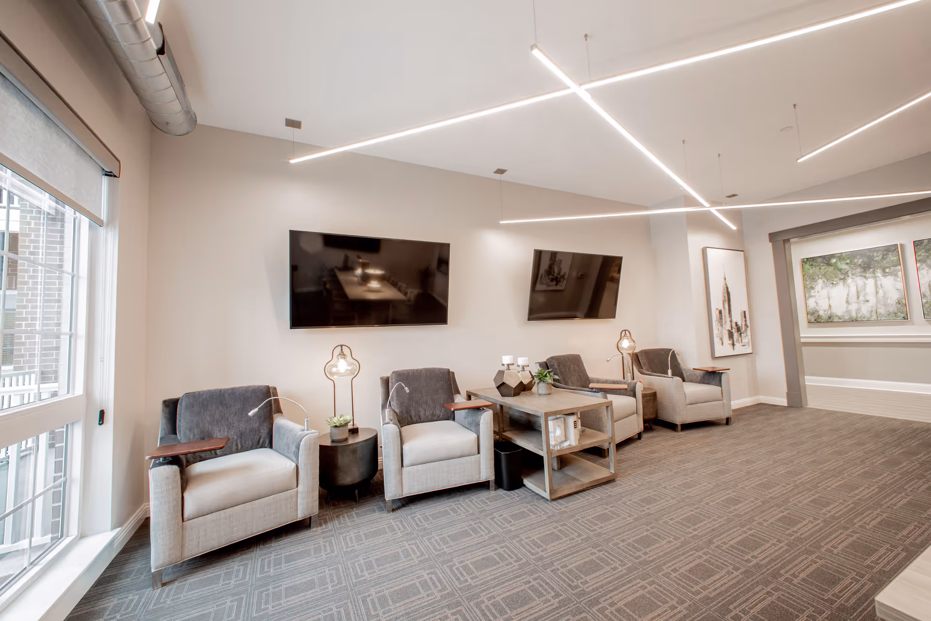 A modern senior living common area with four cushioned armchairs arranged along a wall. Each chair has a small attached wooden table. Between the chairs are two small side tables with decorative items and lamps. Two flat-screen TVs are mounted on the wall above the chairs. The room has large windows with roller shades, contemporary linear ceiling lights, and framed artwork on the walls. The floor is carpeted with a geometric pattern.