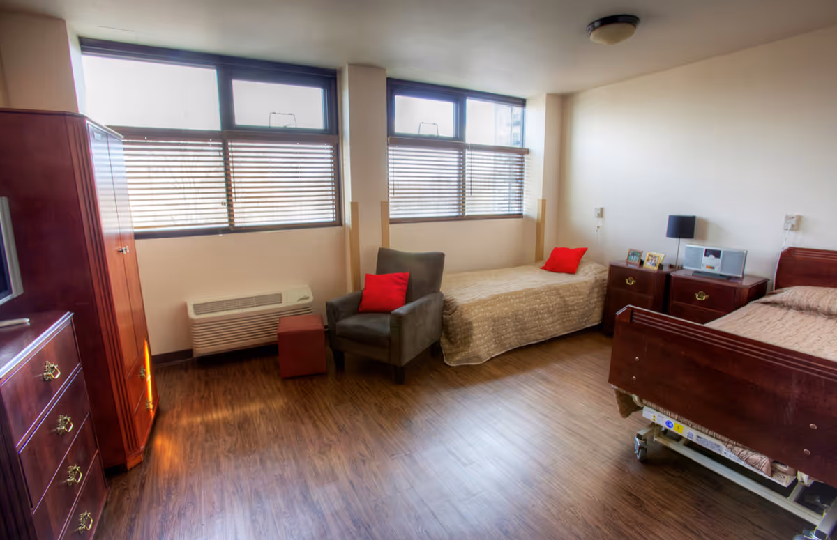 A senior living facility bedroom with two beds, one armchair with a red pillow, wooden furniture including a dresser and nightstands, and large windows with blinds letting in natural light.