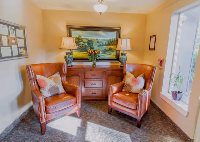 A cozy seating area with two brown leather armchairs, each with a decorative pillow, positioned on either side of a wooden cabinet. On top of the cabinet are two matching table lamps and a vase with flowers. Above the cabinet hangs a framed landscape painting. There is a window to the right with a small potted plant on the windowsill, and a bulletin board on the left wall.