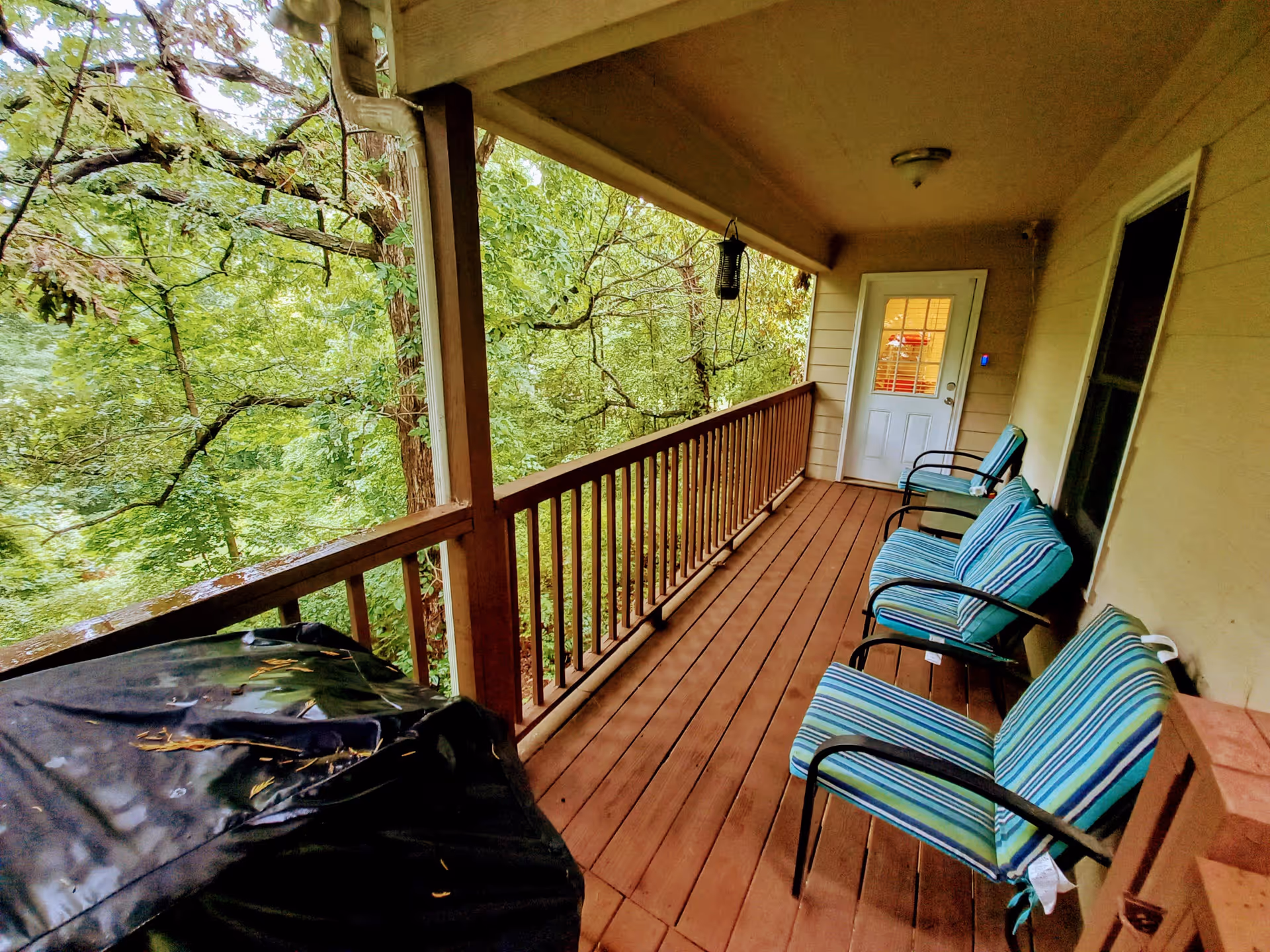 Covered wooden porch with three striped cushioned chairs lined up against the wall, a white door with glass panes at the end, and a black covered grill on the left side. The porch overlooks lush green trees and foliage.