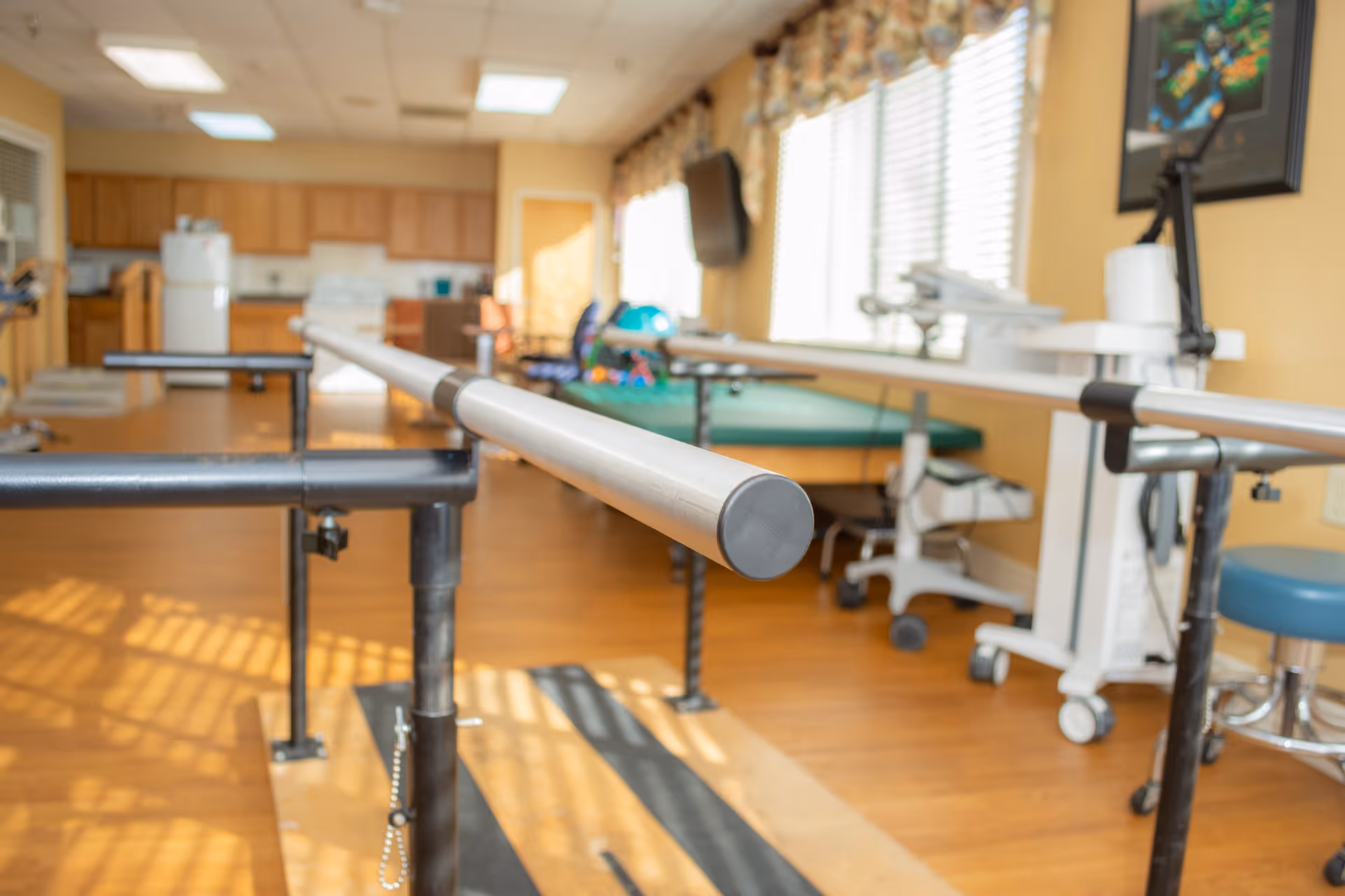 Sunny interior therapy room with parallel walking bars, a treatment table, and medical equipment.