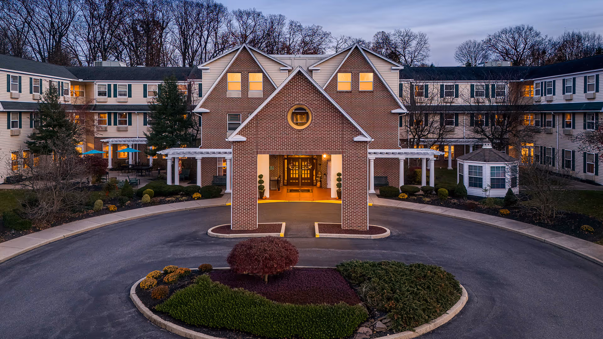 Front exterior view of Atria Bethlehem senior living facility at dusk with a circular driveway, landscaped garden in the center, and warm lights glowing from the building windows.