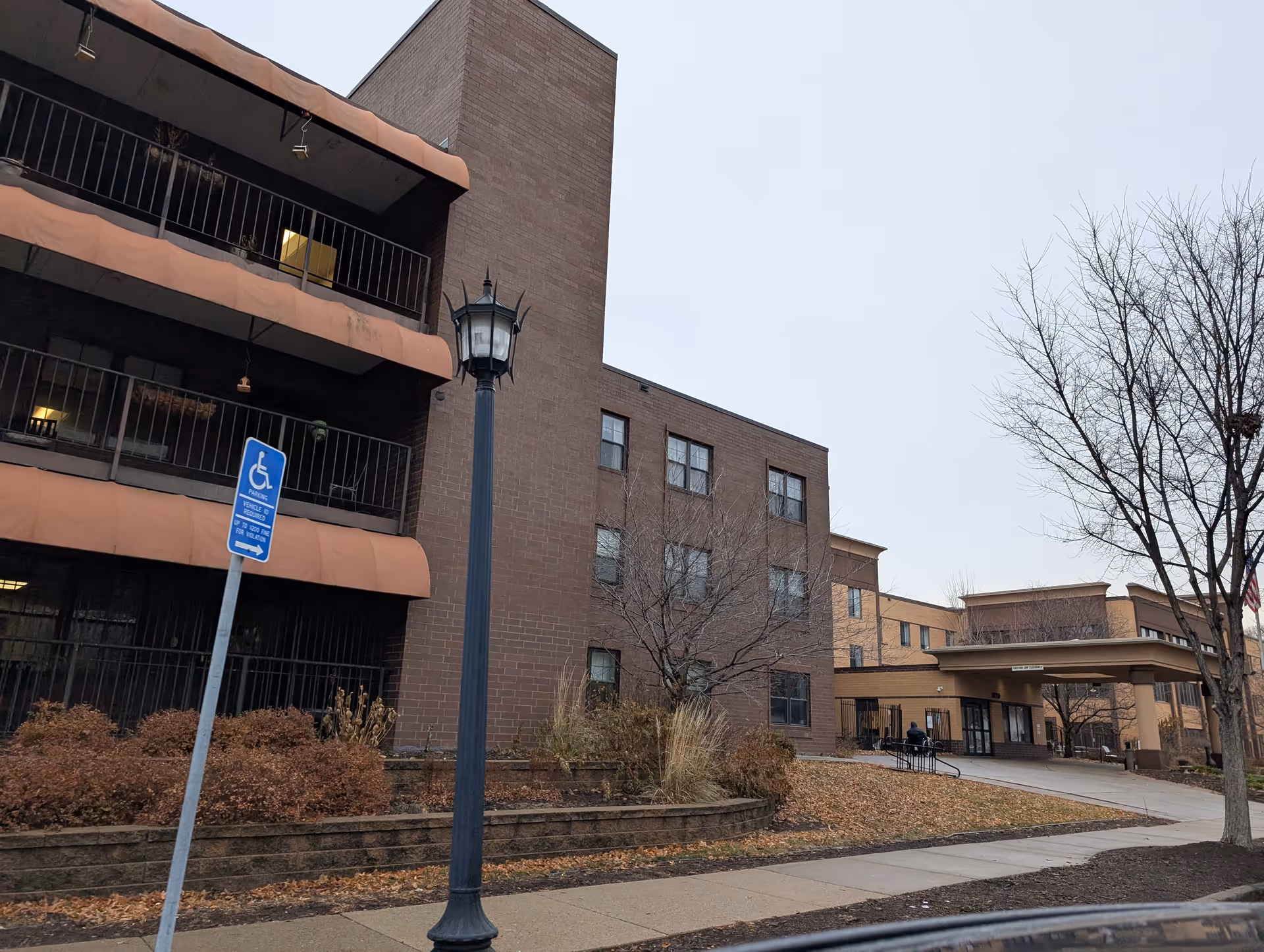 Front exterior of a multi-story brick senior living building with balconies, a covered entrance, lamp post and a handicap parking sign.