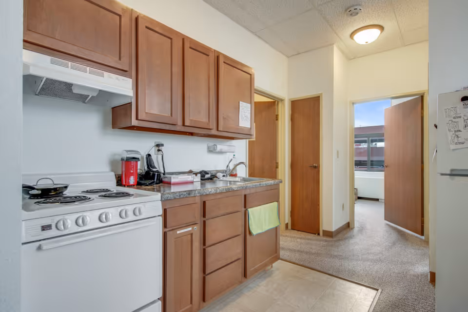 Interior view of a small kitchen area in a senior living facility with wooden cabinets, a white electric stove, a countertop with a sink, a red coffee maker, and a black telephone. There are two wooden doors and an open doorway leading to a carpeted room with a window.