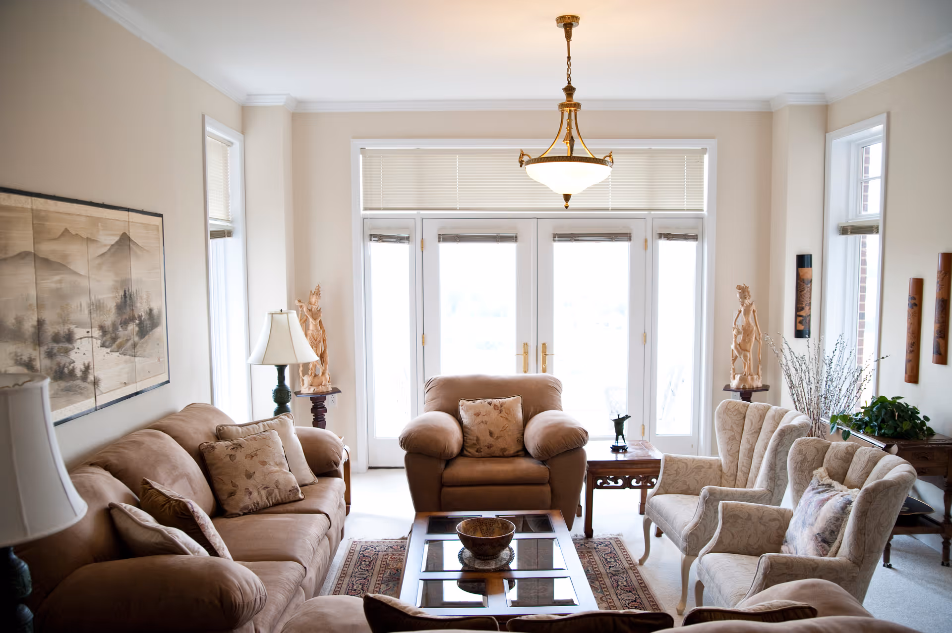 Bright living room with beige sofas and armchairs arranged around a glass coffee table facing large French doors.