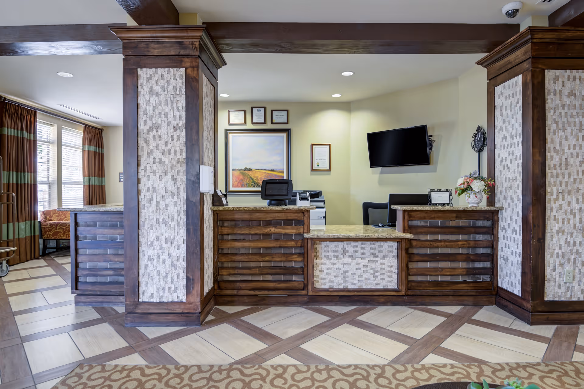 Reception desk area in a senior living facility with wooden paneling and decorative tile accents. Behind the desk are framed certificates and a landscape painting on the wall, along with a mounted flat-screen TV. To the left, there is a window with brown and green striped curtains and a glimpse of a seating area.