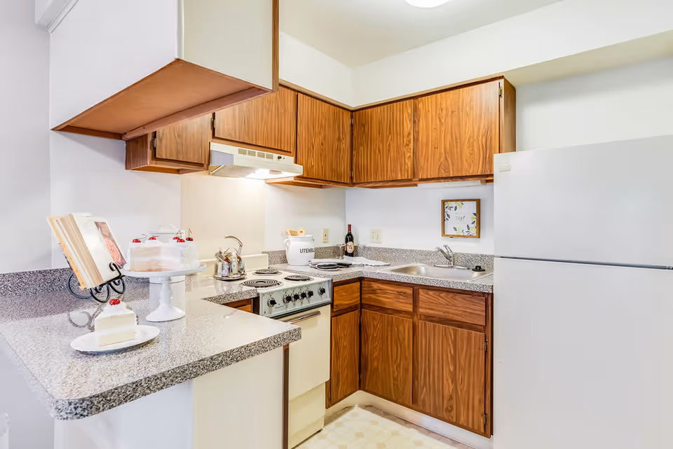A compact kitchen with wooden cabinets, a white refrigerator, an electric stove with four burners, a stainless steel sink, and a speckled gray countertop. On the counter, there is a slice of cake on a plate, a cake stand with a cake topped with cherries, a cookbook on a stand, a metal kettle on the stove, and a container labeled 'UTENSILS'.